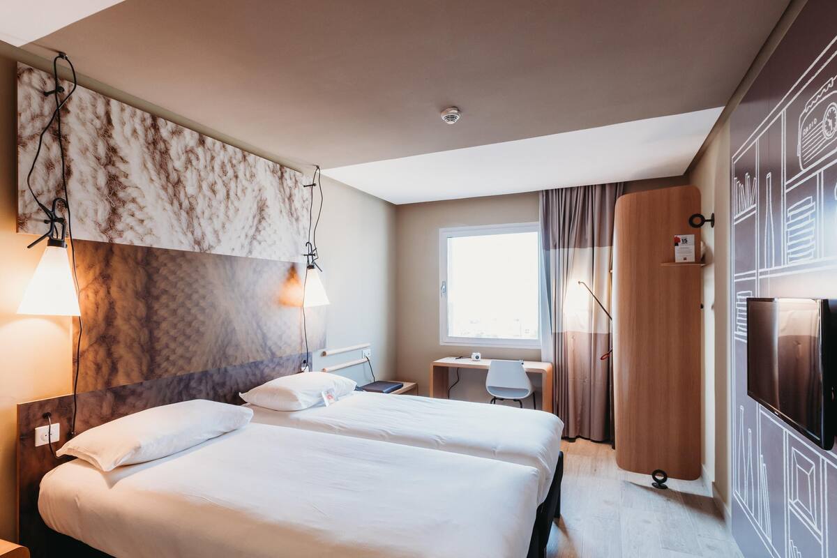 Una habitación de hotel con dos camas, un escritorio, una silla, un armario y un ventanal grande.