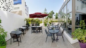 Un espace extérieur aménagé en terrasse, avec des tables et des chaises, entouré de plantes en pot et agrémenté d’un parasol rouge.