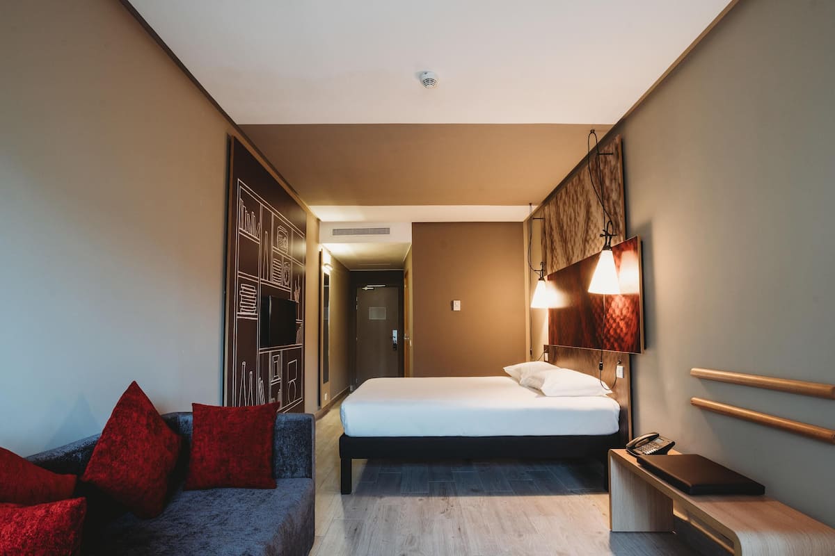 Habitación de hotel con cama, escritorio con teléfono, sofá con cojines rojos y televisor montado en la pared.