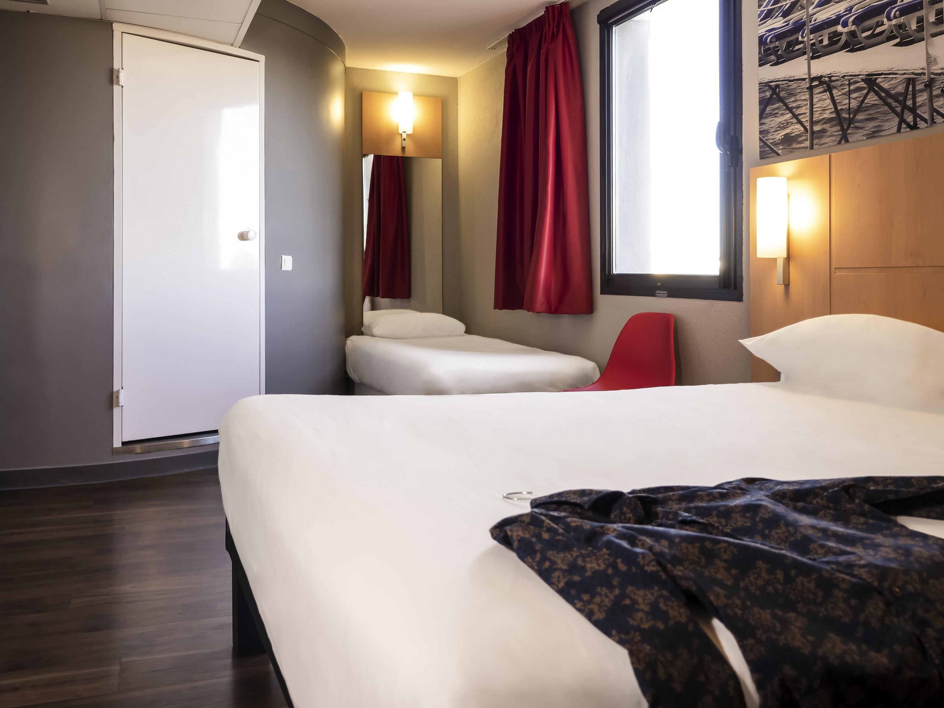 Une chambre d’hôtel avec un lit, une chaise rouge, une petite table et une fenêtre avec des rideaux.