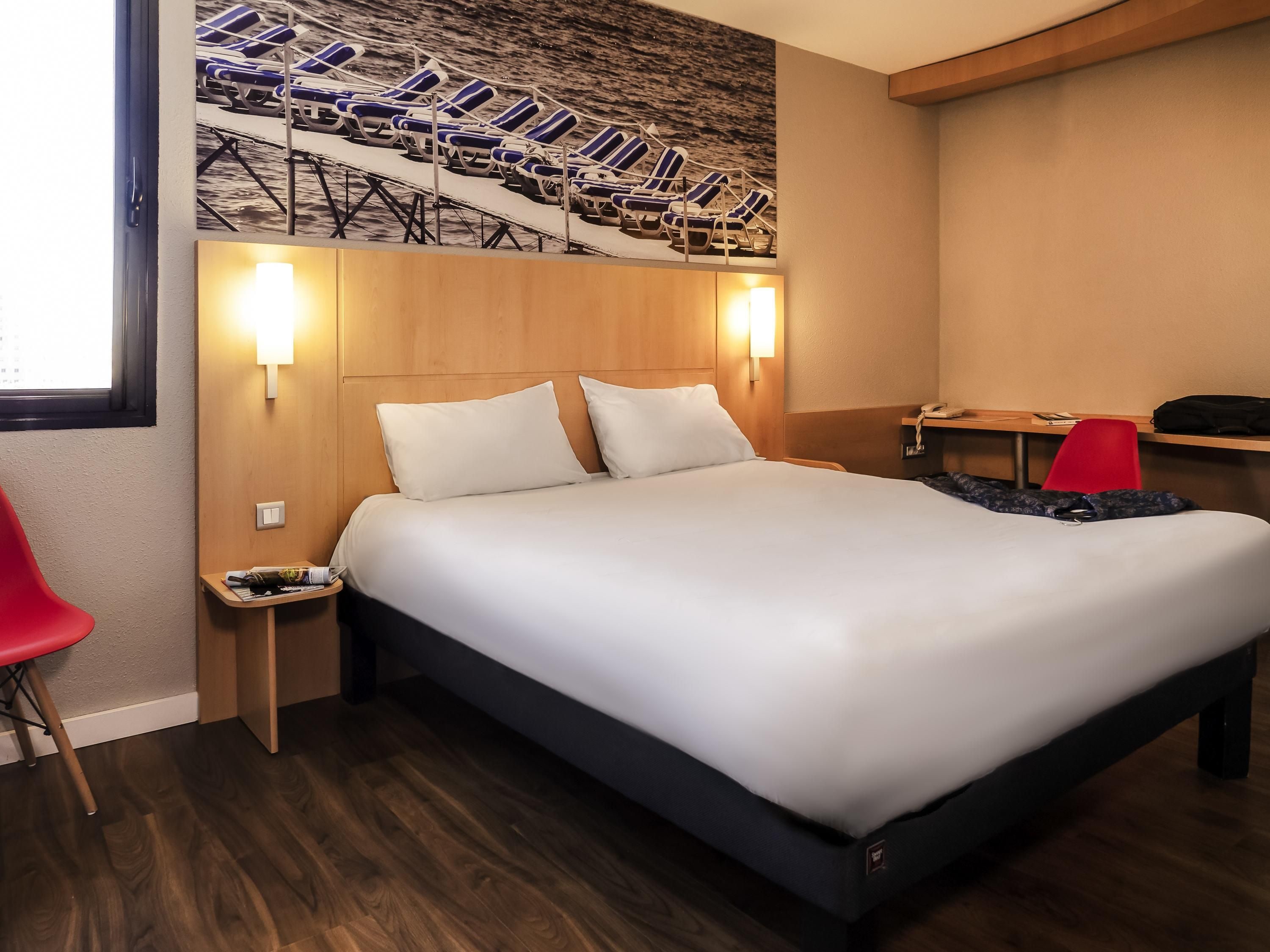 A hotel room with a large bed, two bedside tables, a red chair, a desk with a telephone, and a wall-mounted artwork of lounge chairs.