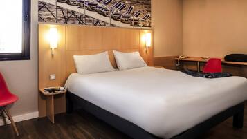 A hotel room with a large bed, two bedside tables, a red chair, a desk with a telephone, and a wall-mounted artwork of lounge chairs.