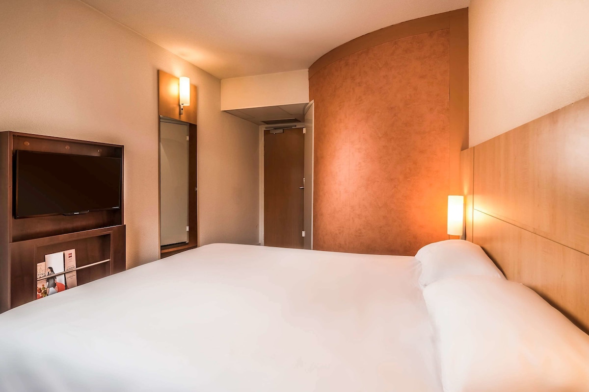 A hotel room with a large bed, a flat-screen TV, a wardrobe, and a mirror.