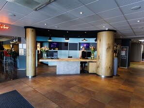 A reception area with a wooden reception desk, a digital screen, and a seating area with chairs and a plant. - ibis Cannes Mandelieu (Mandelieu-La-Napoule)