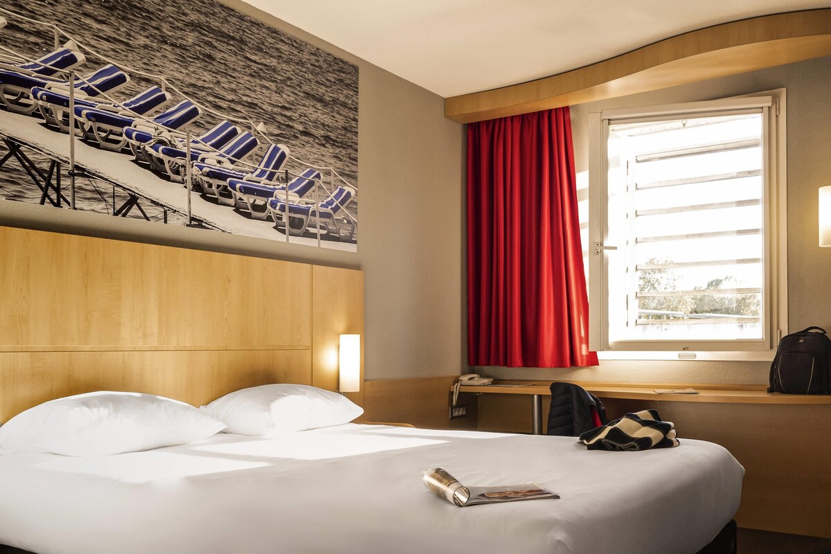 A hotel room with a bed, a desk, a chair, a window with red curtains, and a wall mural of beach chairs.