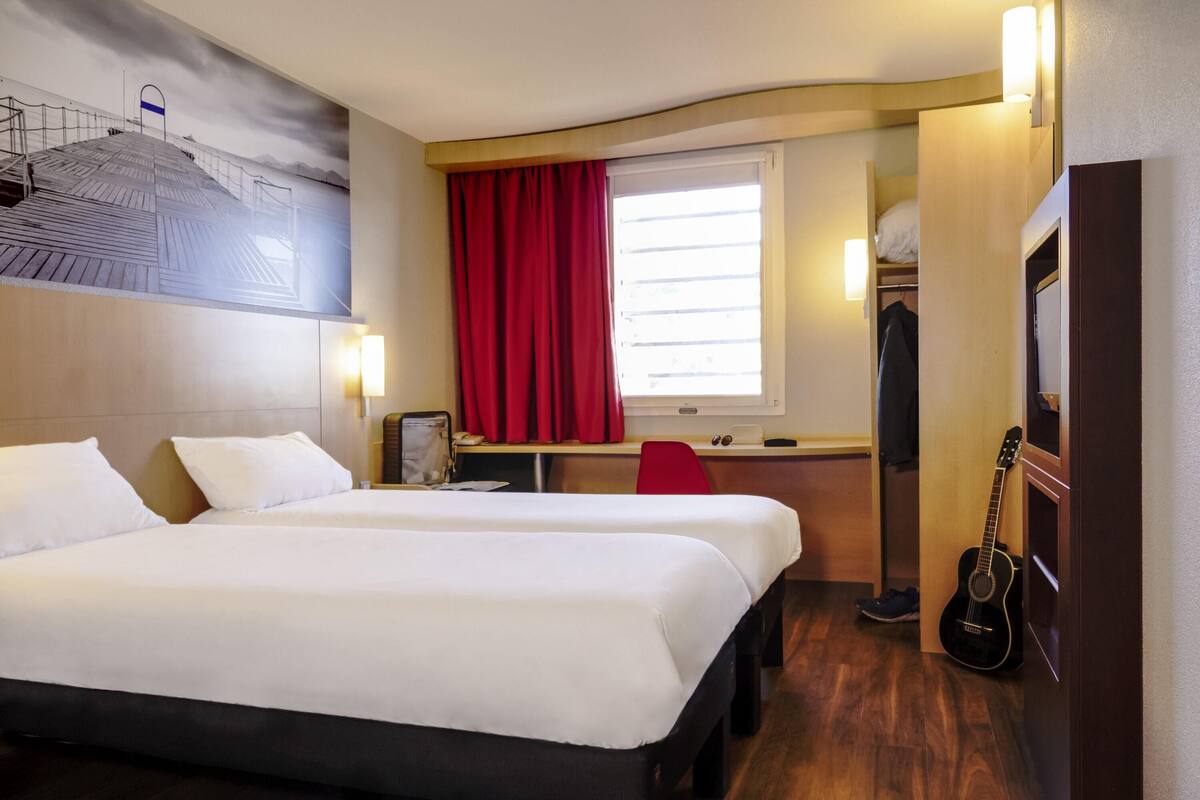A hotel room with two beds, a desk, a chair, a guitar, and a window with red curtains.