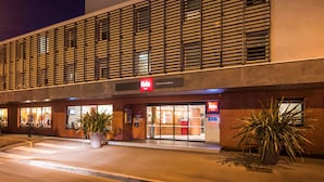 Um hotel Ibis moderno com uma placa vermelha grande, amplas janelas e plantas em vasos à frente.