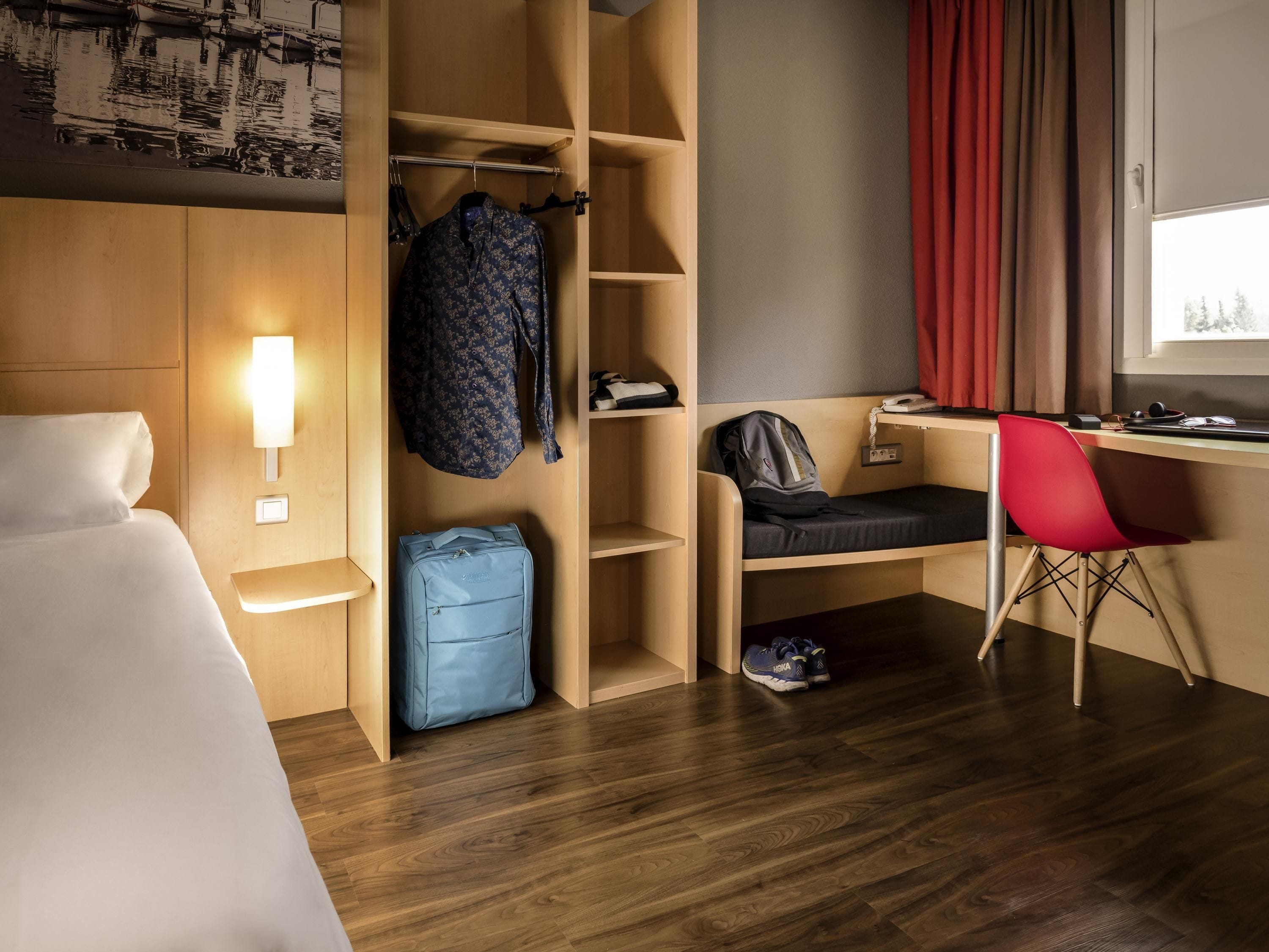 A hotel room with a bed, a desk with a chair, a wardrobe, and a suitcase.