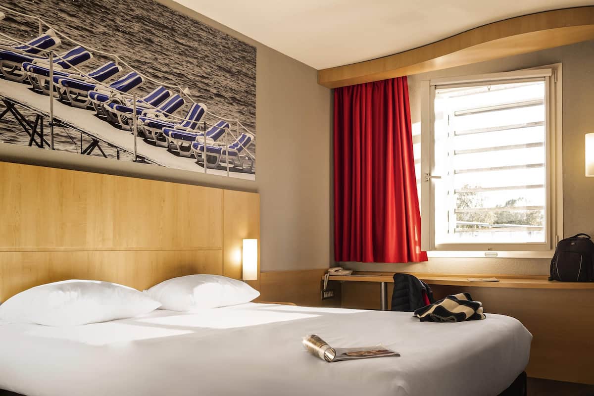 A hotel room with a bed, a desk, a chair, a window with red curtains, and a wall mural of beach chairs.