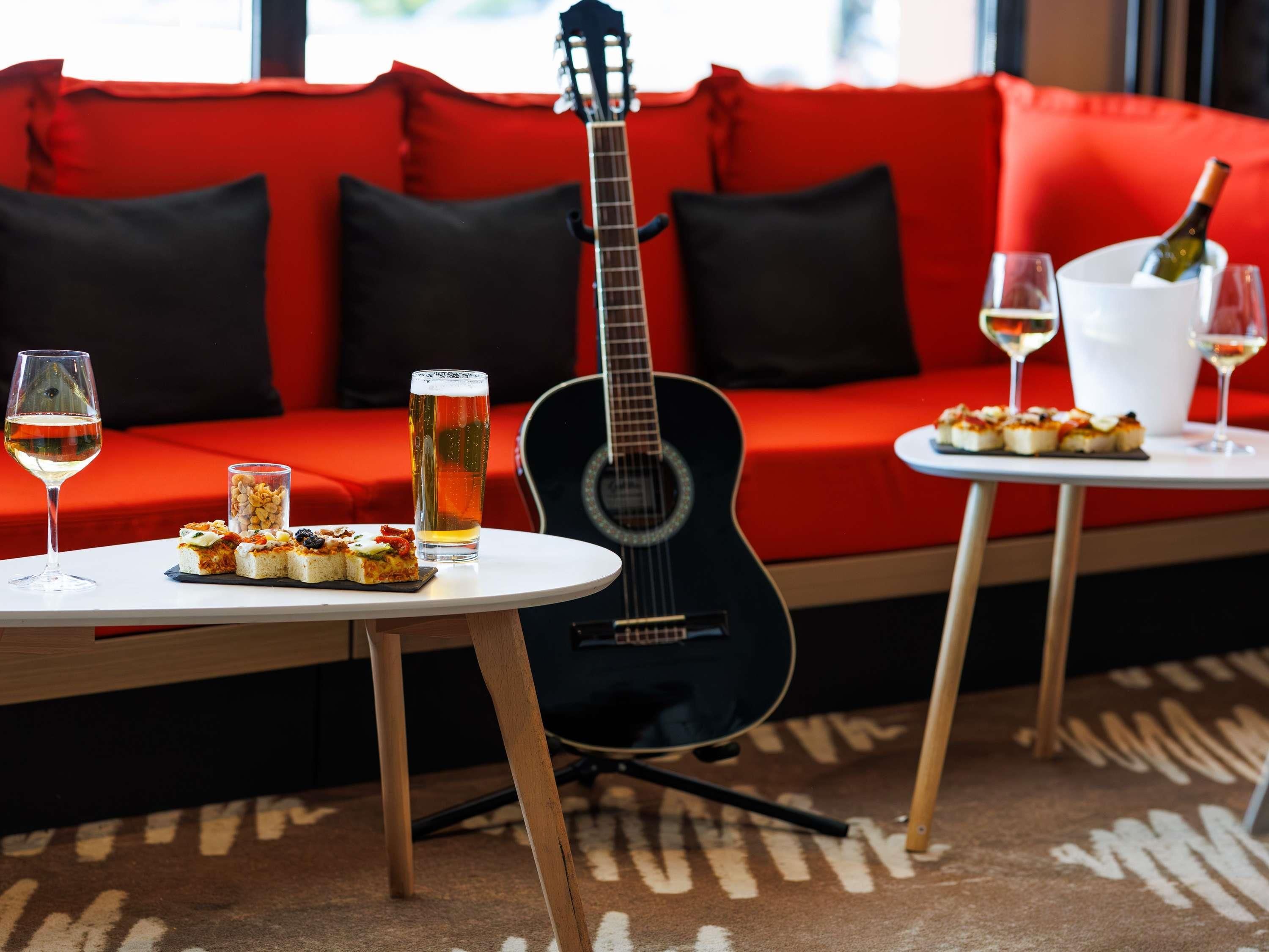 Un sofá rojo con cojines negros, una guitarra, una mesa con una jarra de cerveza, una copa de vino y una botella de champán.