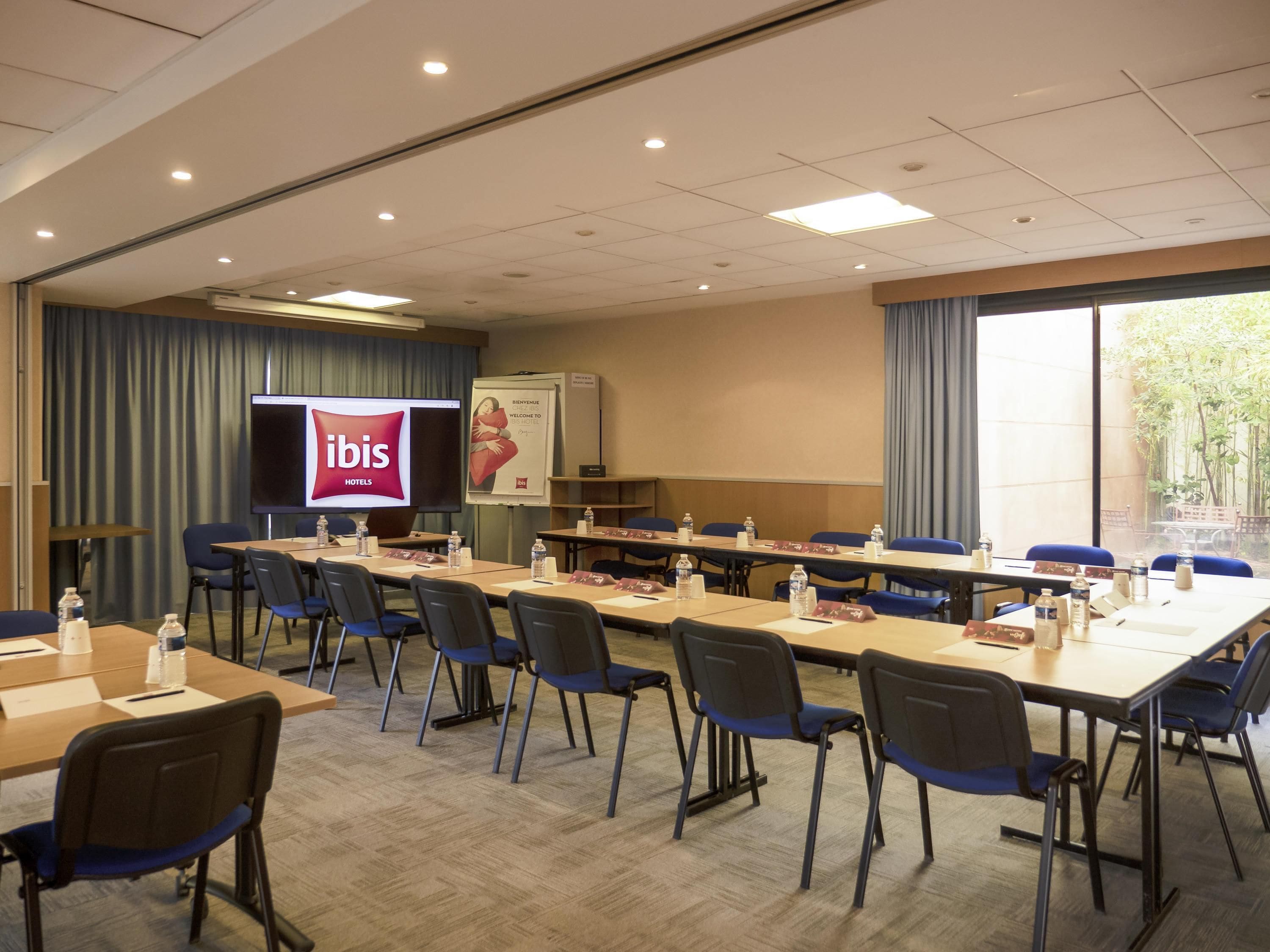 Een vergaderruimte met een groot scherm waarop 'ibis Hotels' staat, een whiteboard en een uitzicht op het omliggende landschap door grote ramen.