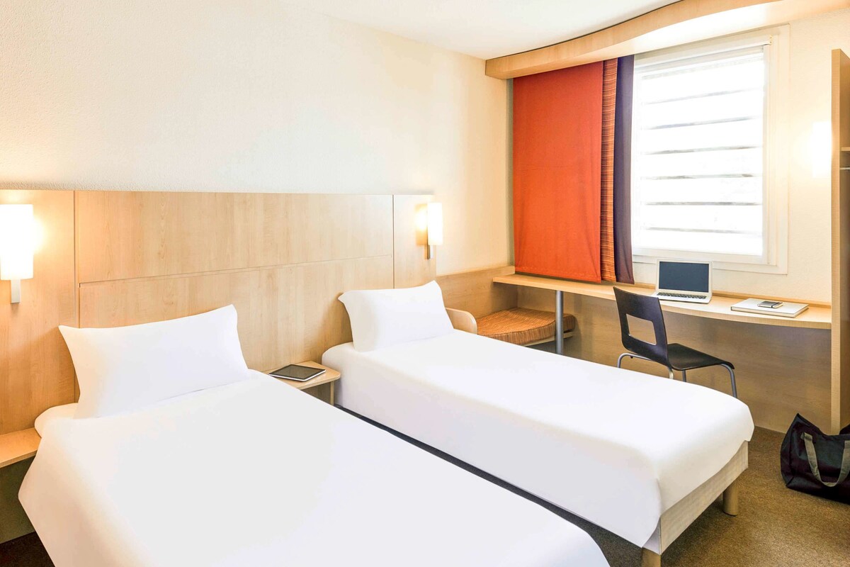 A hotel room with two single beds, a desk with a laptop, a chair, and a window with curtains.