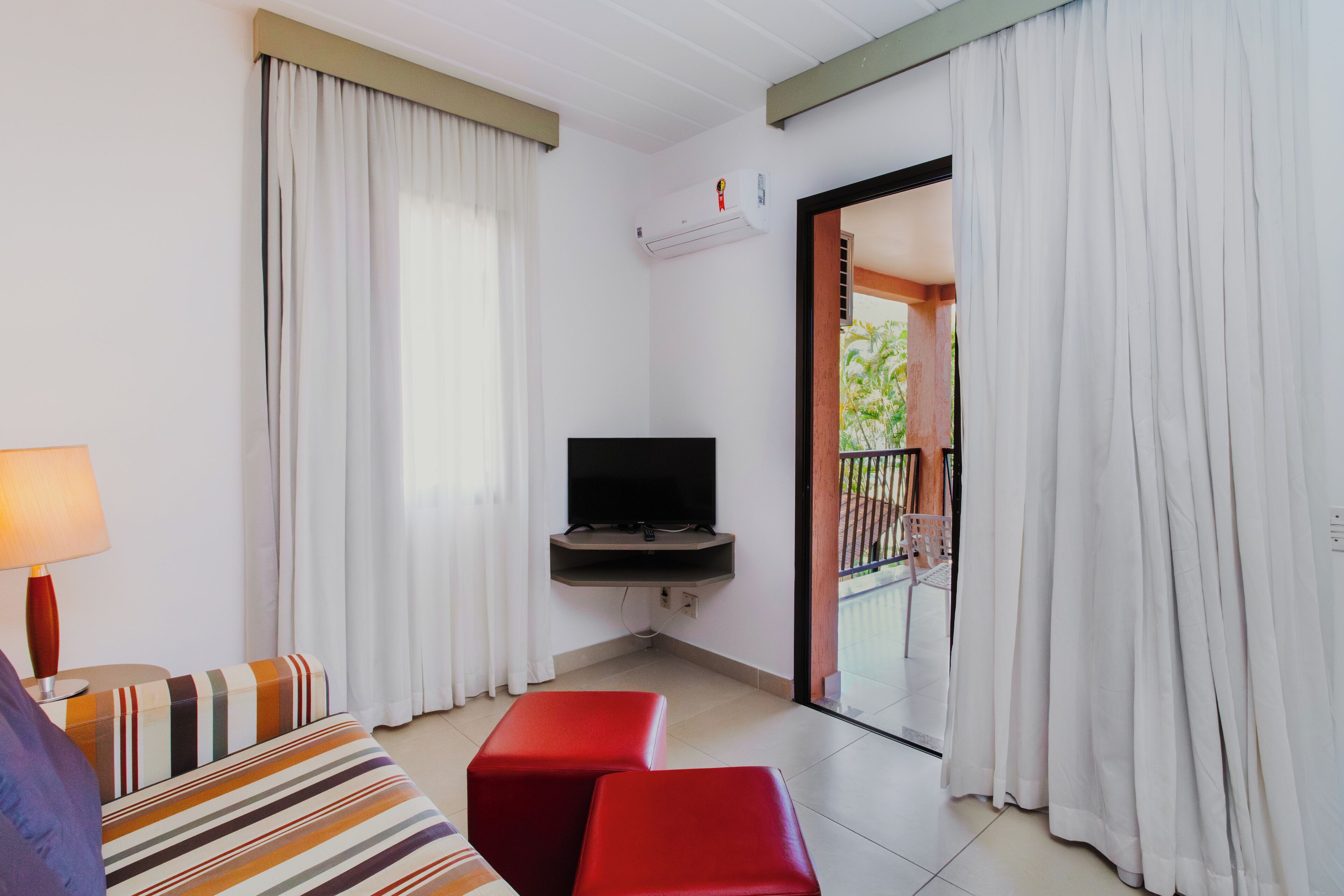 A hotel room with a TV, a sofa, a red ottoman, and a balcony with curtains.