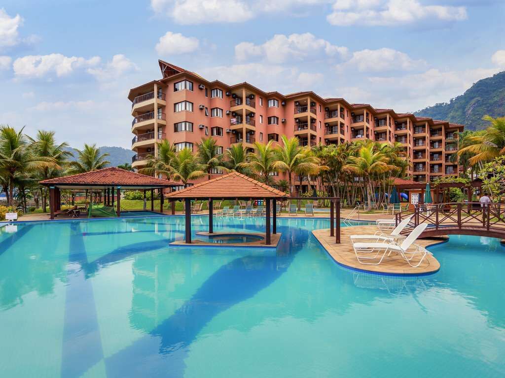 Un hotel con una piscina grande, palmeras y montañas al fondo.