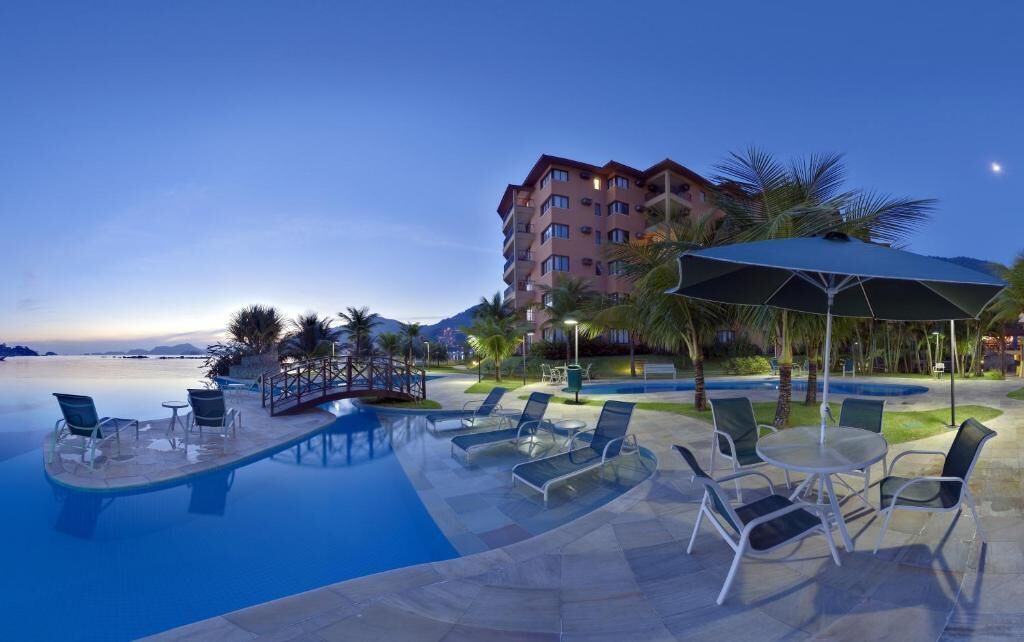 Ett hotellområde med en pool, solstolar och parasoller, omgivet av palmer och en byggnad med flera våningar.