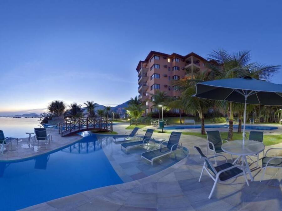 Zona de piscina de hotel con sillas reclinables y sombrillas, rodeada de palmeras y un edificio de varios pisos.