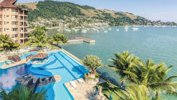 A resort with a swimming pool, palm trees, and a view of the sea and hills.