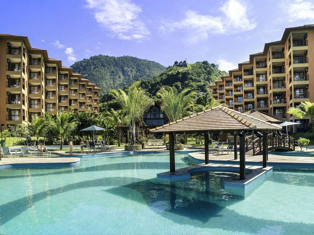 A resort pool area with a gazebo, palm trees, and a mountainous backdrop.