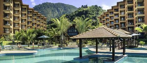 A resort pool area with a gazebo, palm trees, and a mountainous backdrop.