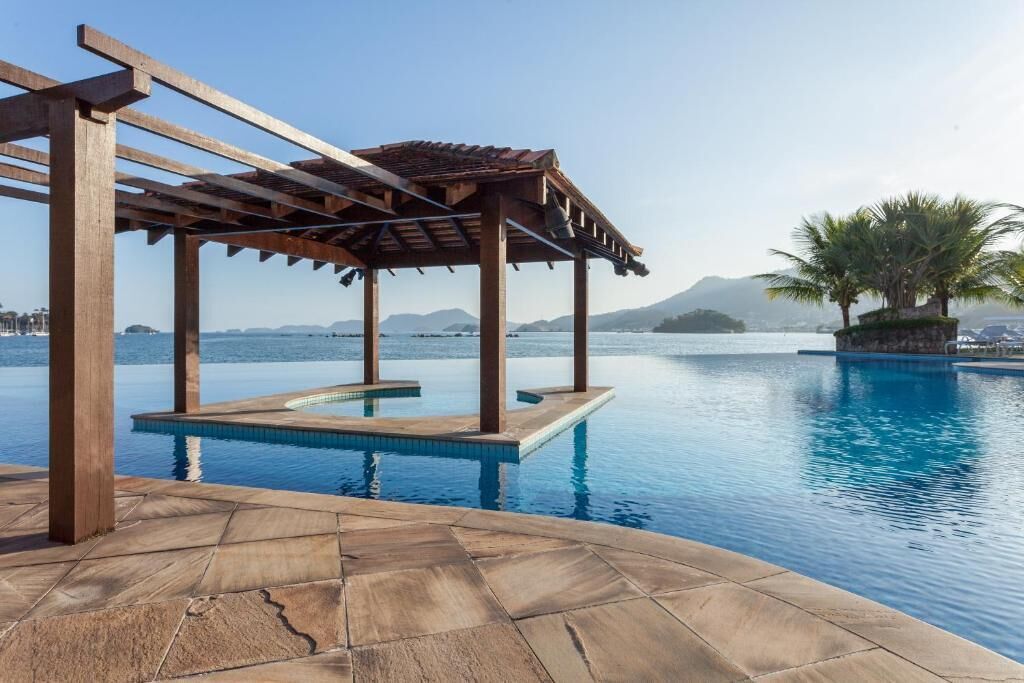 A wooden pergola over a swimming pool with a clear view of the sea and mountains in the background.
