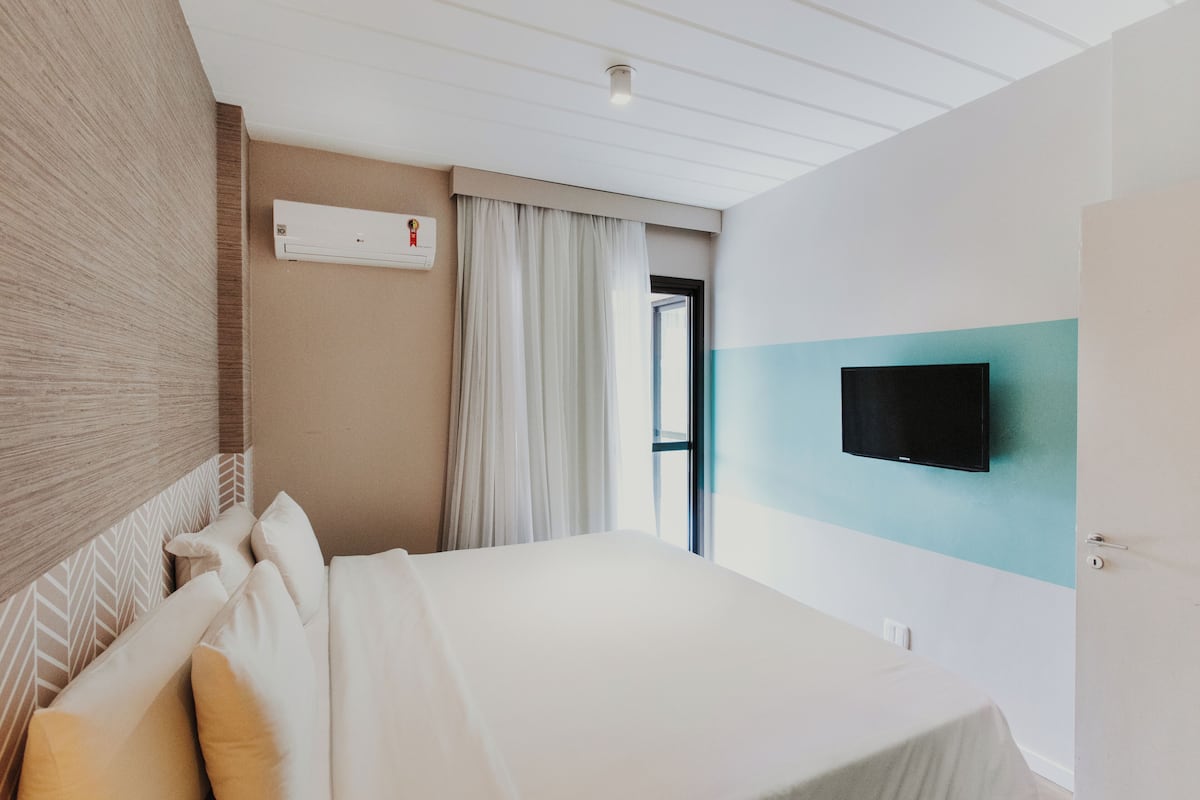 A hotel room with a bed, a flat-screen TV mounted on the wall, a window with curtains, and an air conditioning unit.
