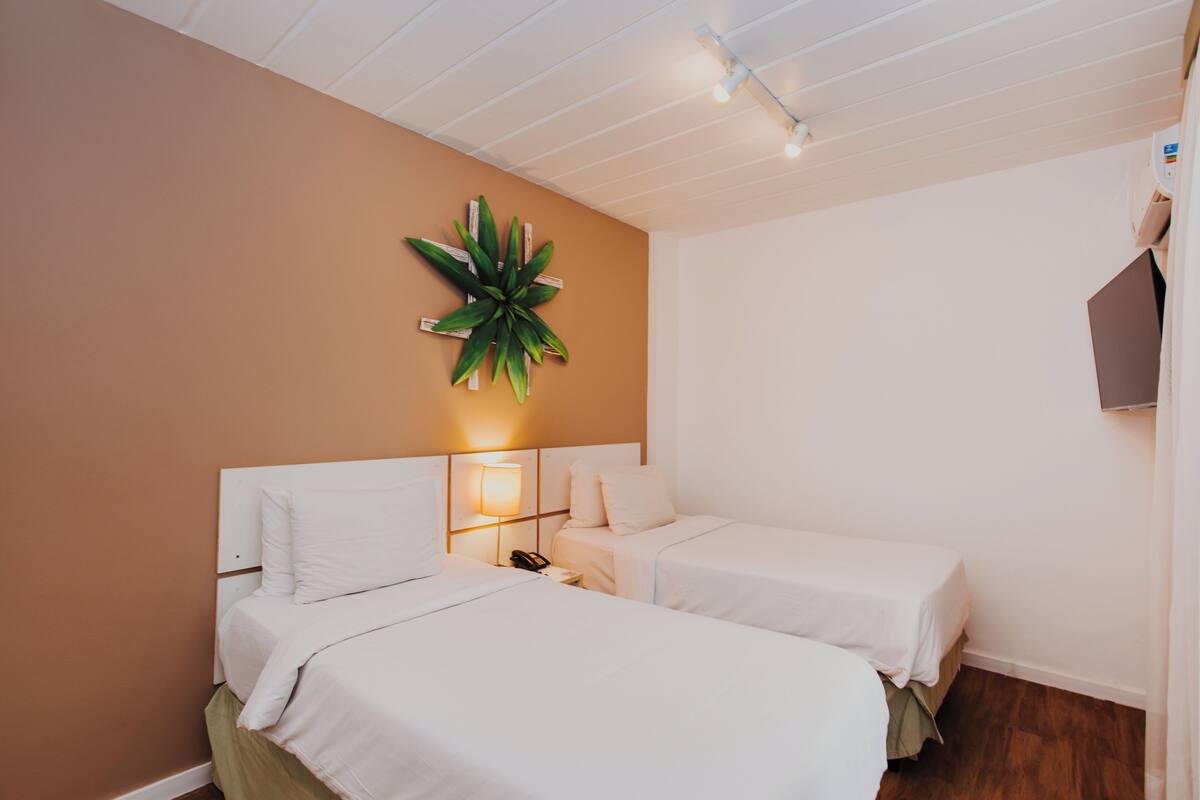 A hotel room with two beds, a wall-mounted plant, and a bedside table with a lamp.