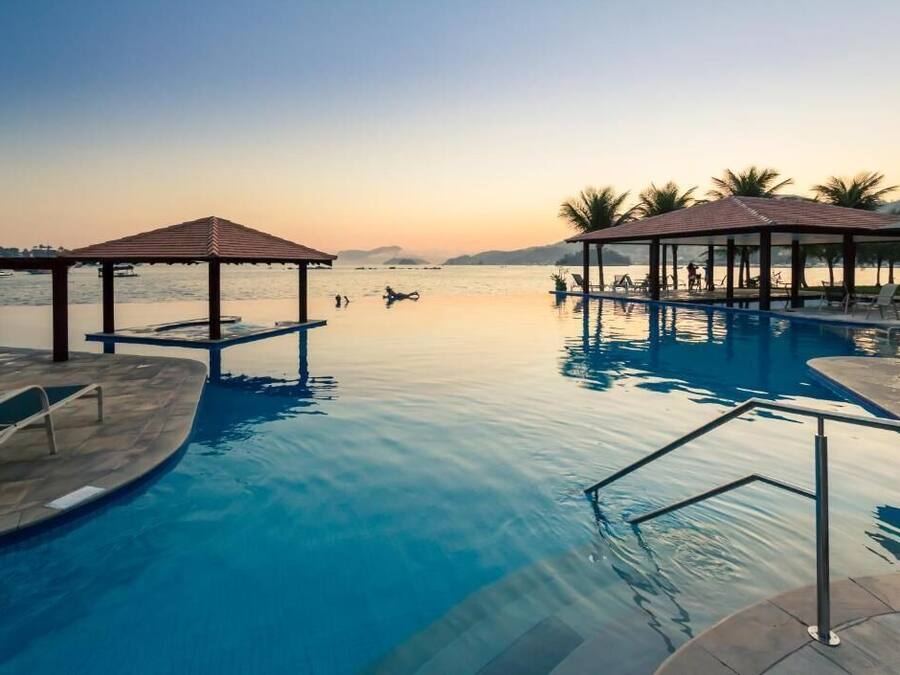 A serene pool area with clear water, two covered gazebos, and a calm sunset in the background.