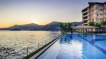 Une piscine avec vue sur la mer et les montagnes, un espace détente couvert et un immeuble à plusieurs étages.
