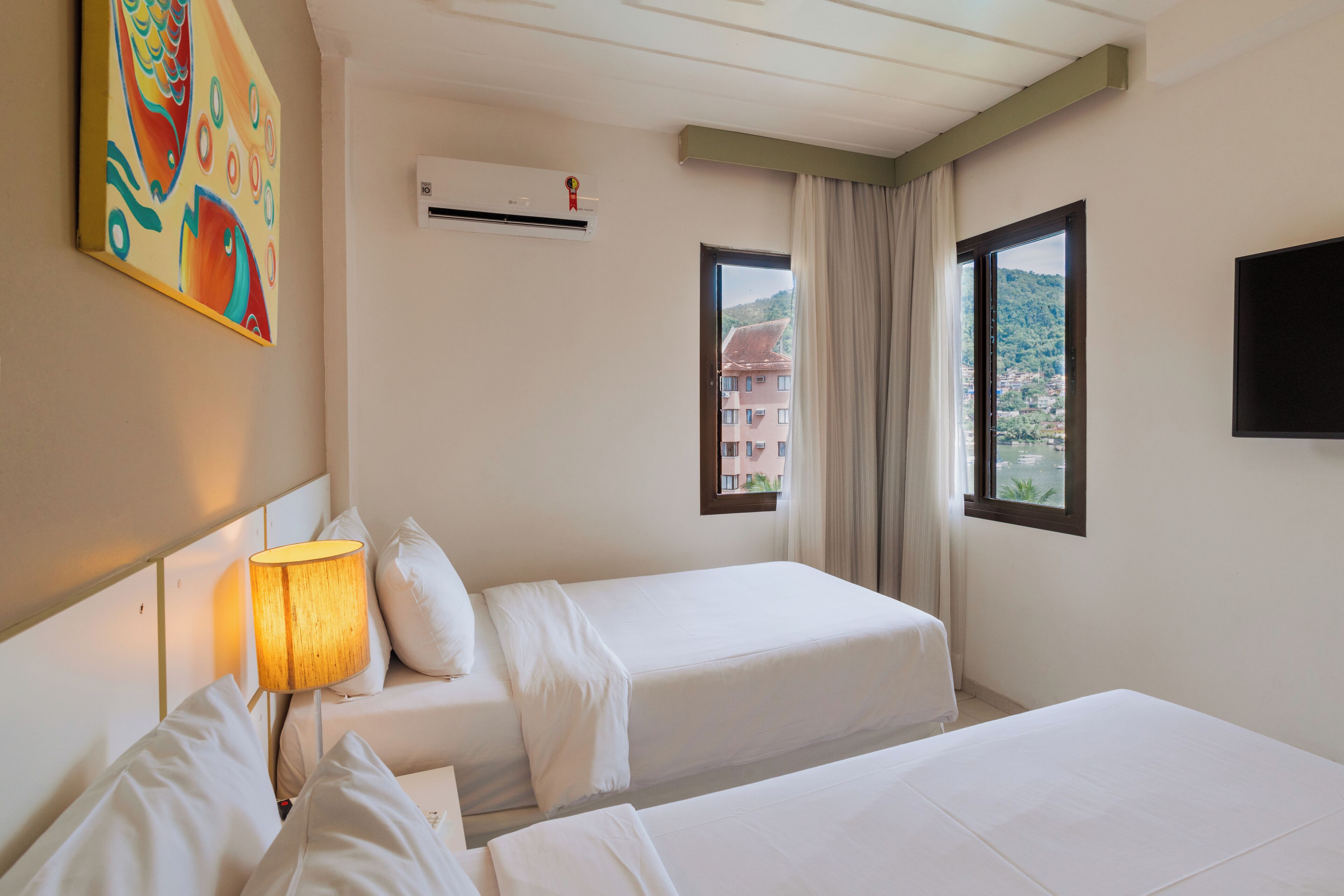 A hotel room with two beds, a lamp, an air conditioner, a TV, and a painting on the wall.