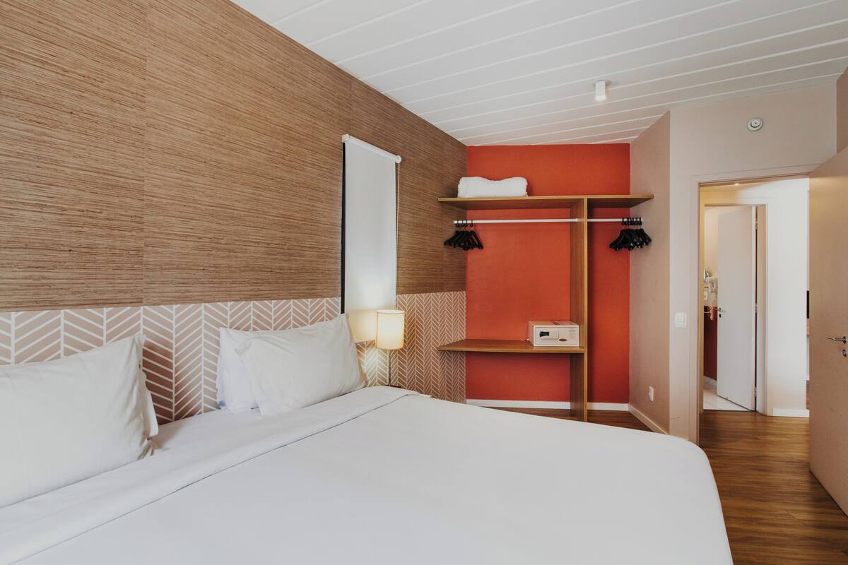 A modern hotel room with a large bed, wooden headboard, a red accent wall, and a built-in shelf with hanging clothes.