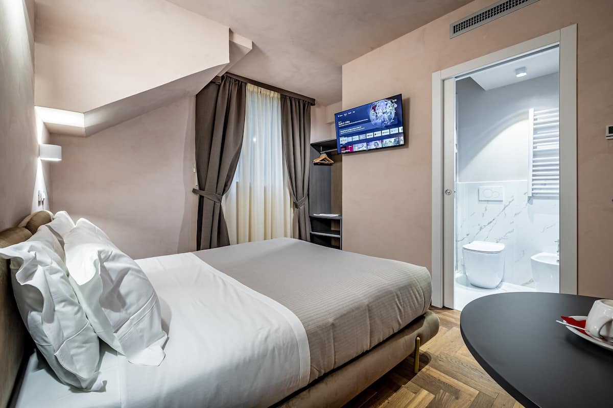 A hotel room with a bed, a TV mounted on the wall, a bathroom with a toilet and sink, and a small table with a cup.