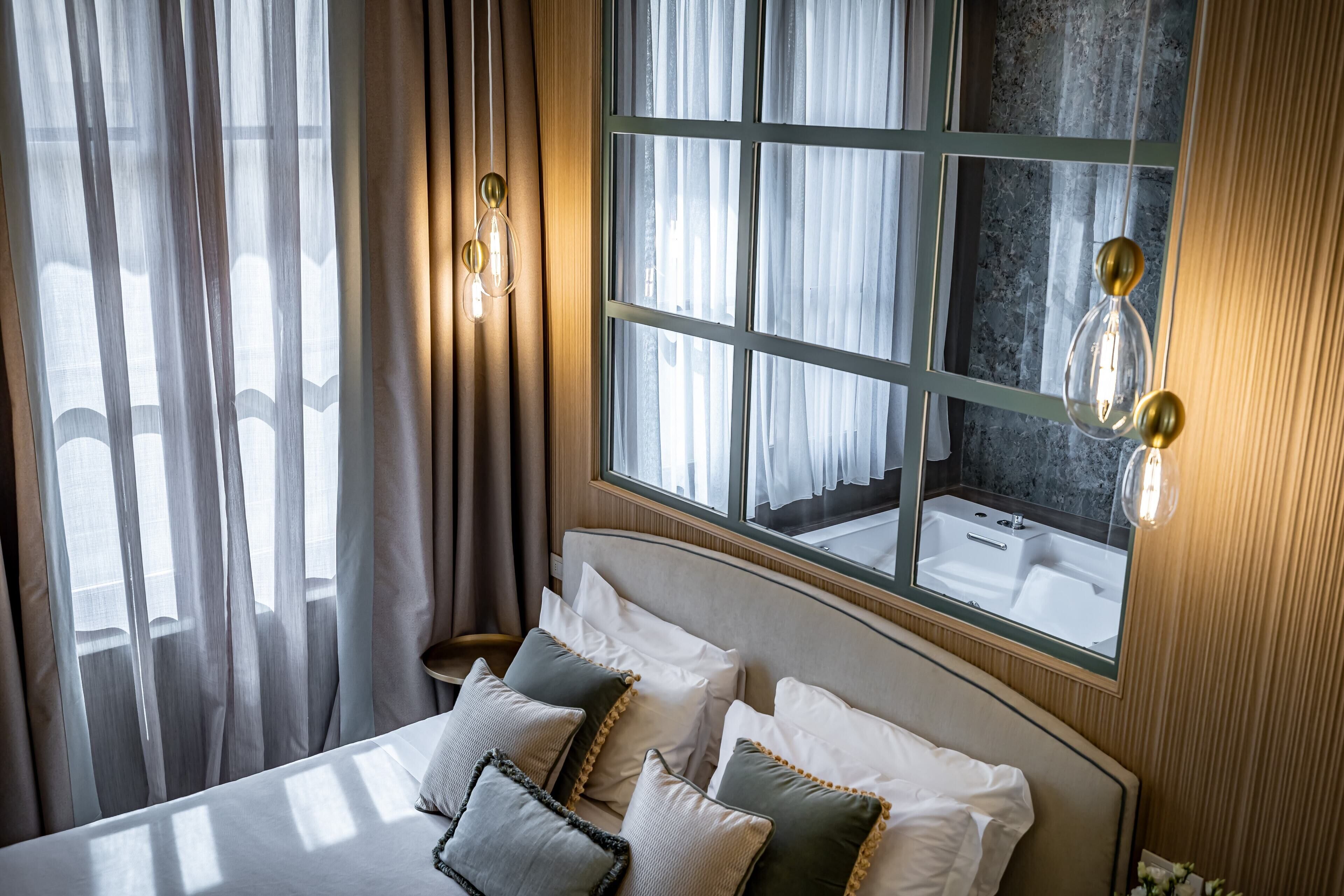 A hotel room with a large bed, a view of a bathroom through a window, and a wall-mounted light fixture.