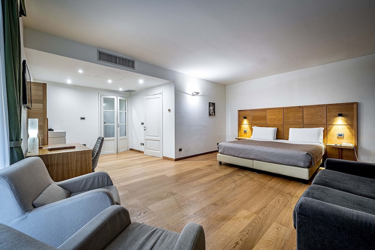 A modern hotel room with a wooden floor, a bed with a wooden headboard, a desk with a chair, and a wardrobe.