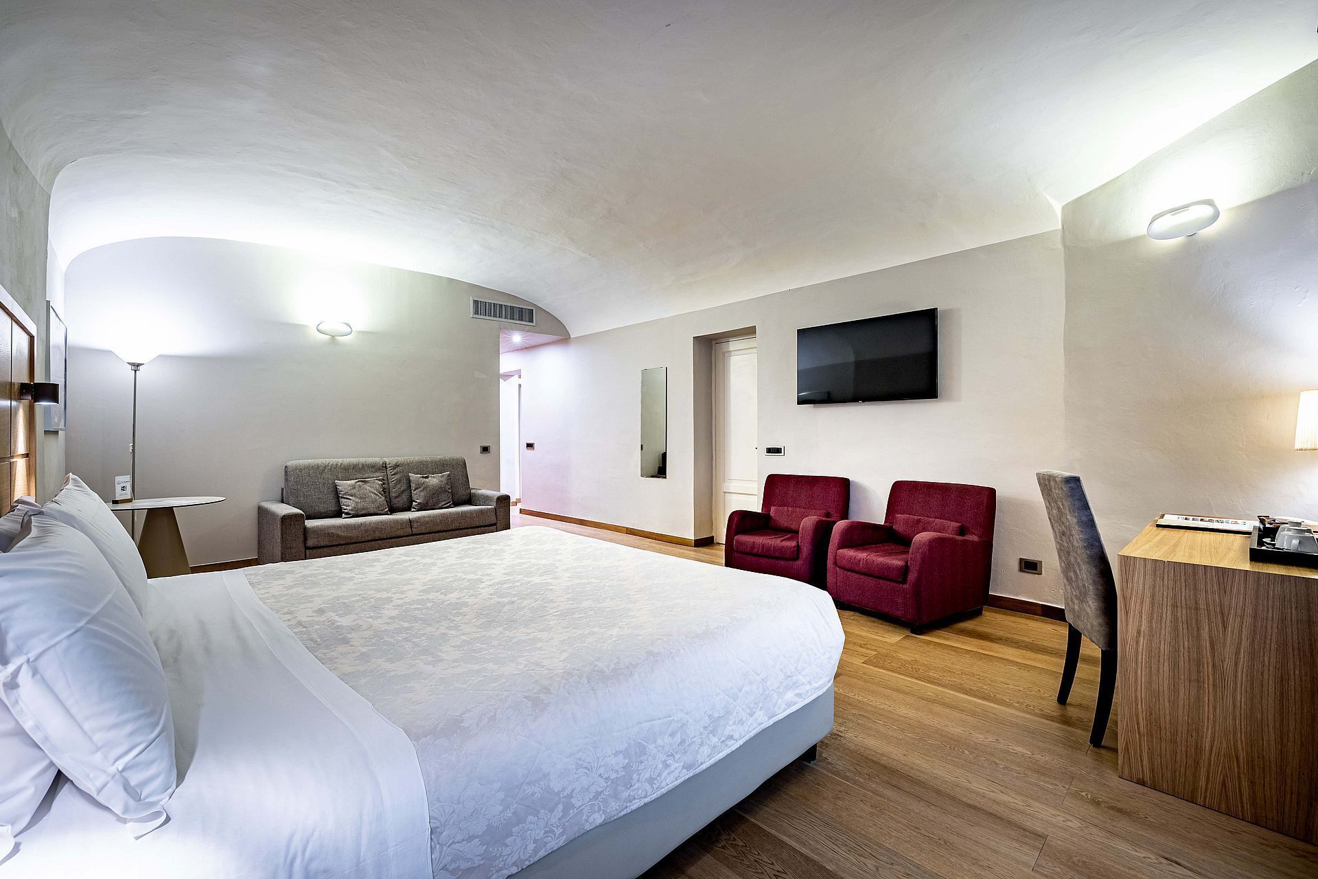 Quarto de hotel com uma cama grande, uma escrivaninha, duas poltronas vermelhas, uma TV fixada na parede e uma mesa de cabeceira de madeira.