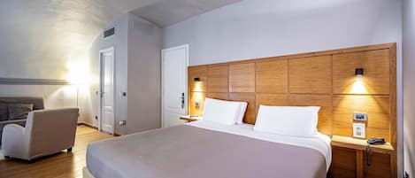 A modern hotel room with a large bed, a wooden headboard, bedside tables with lamps, a sofa, and a door with a vent.