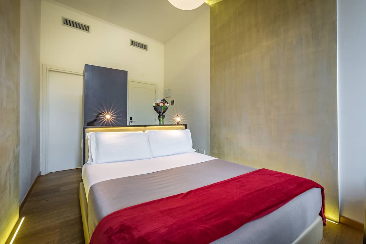 A modern hotel room with a large bed, a red throw blanket, a mirror above the bed, and a bouquet of flowers on the nightstand.