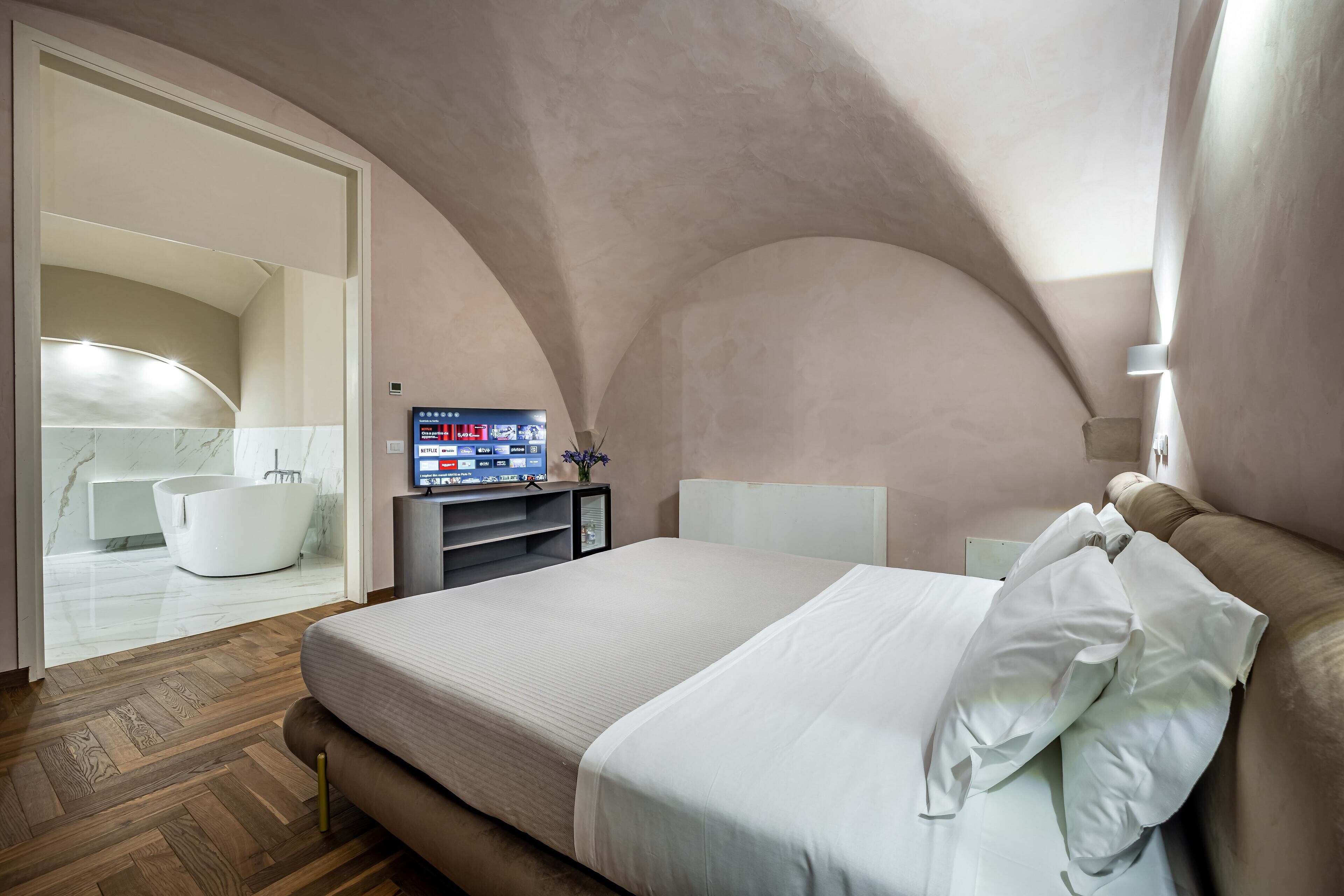 A bedroom with a bed, a TV, a bathroom with a bathtub, and a wooden floor.