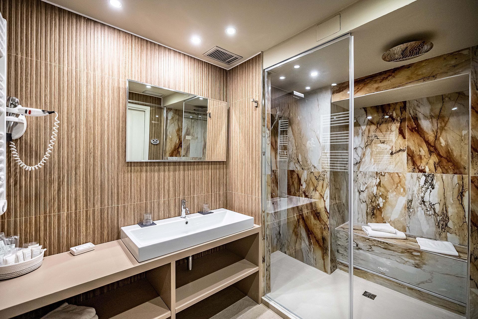 Un baño moderno con tocador de madera, un espejo grande, cabina de ducha de vidrio y paredes de mármol.