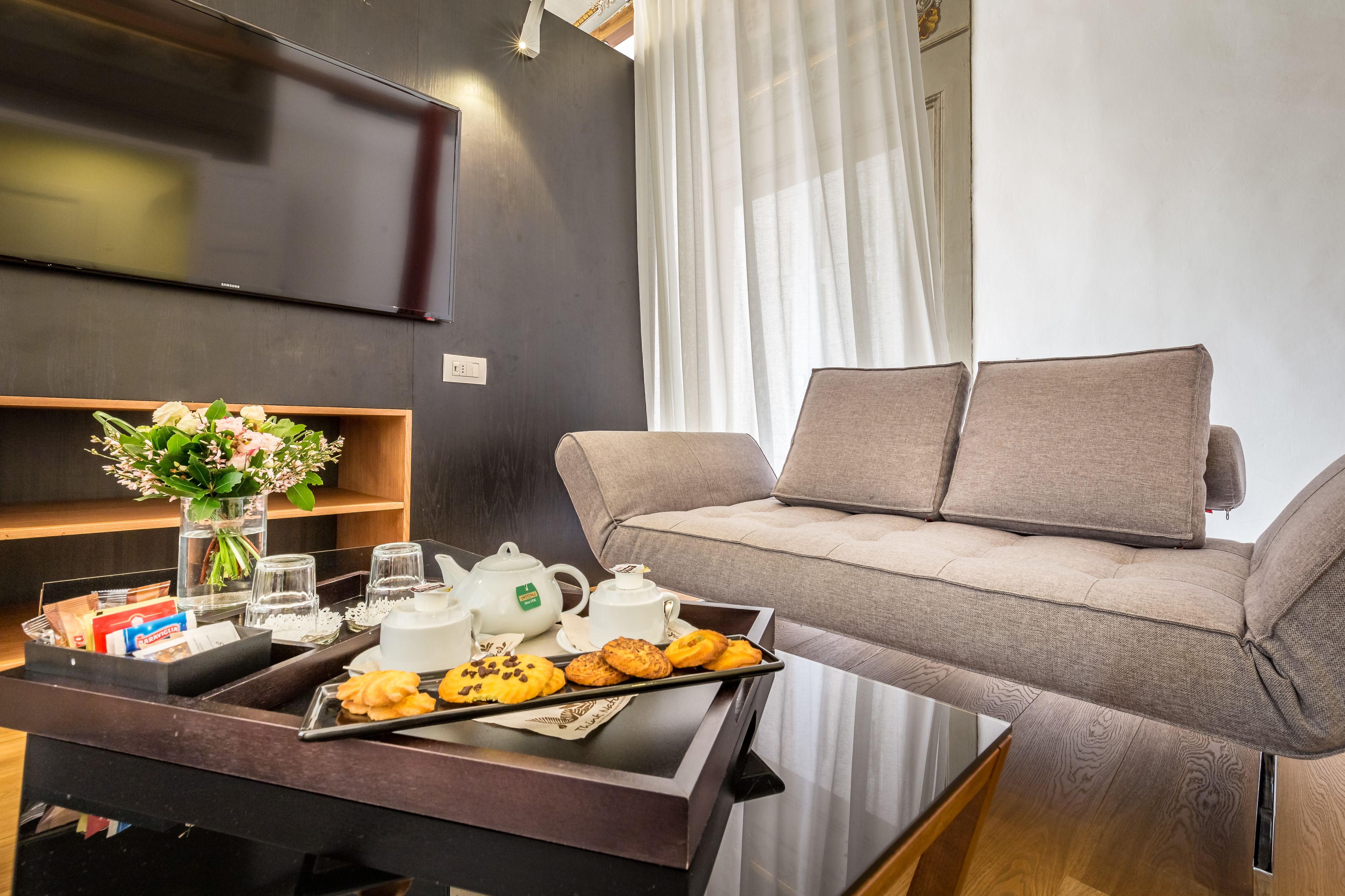 Una sala de estar moderna con un sofá, una mesita con galletas y té, un televisor y un florero con flores.