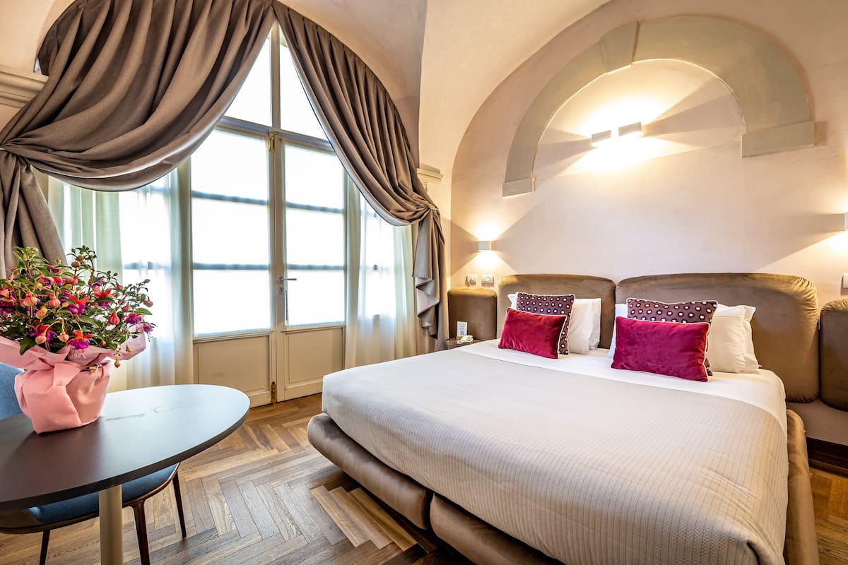 A hotel room with a large bed, a round table with a vase of flowers, and a window with curtains.