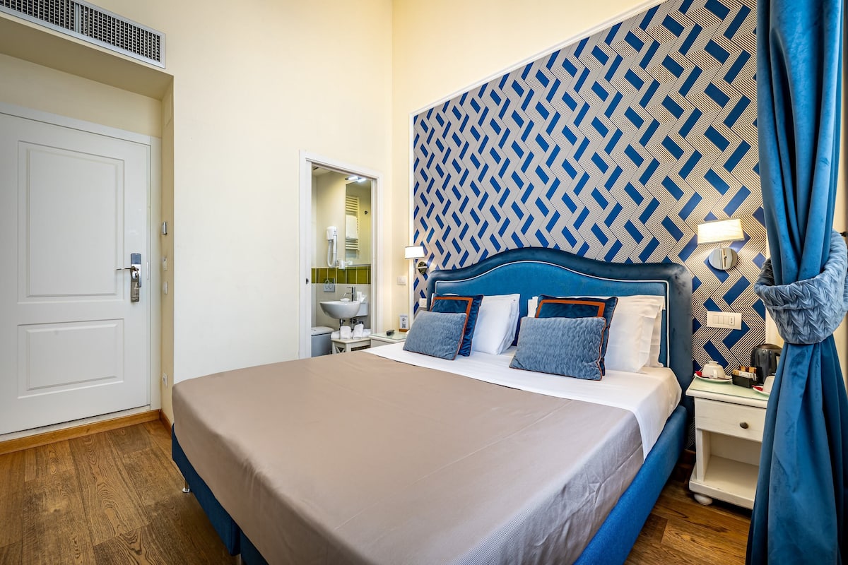 A hotel room with a large bed, a bedside table, a nightstand, a mirror, and a patterned headboard.