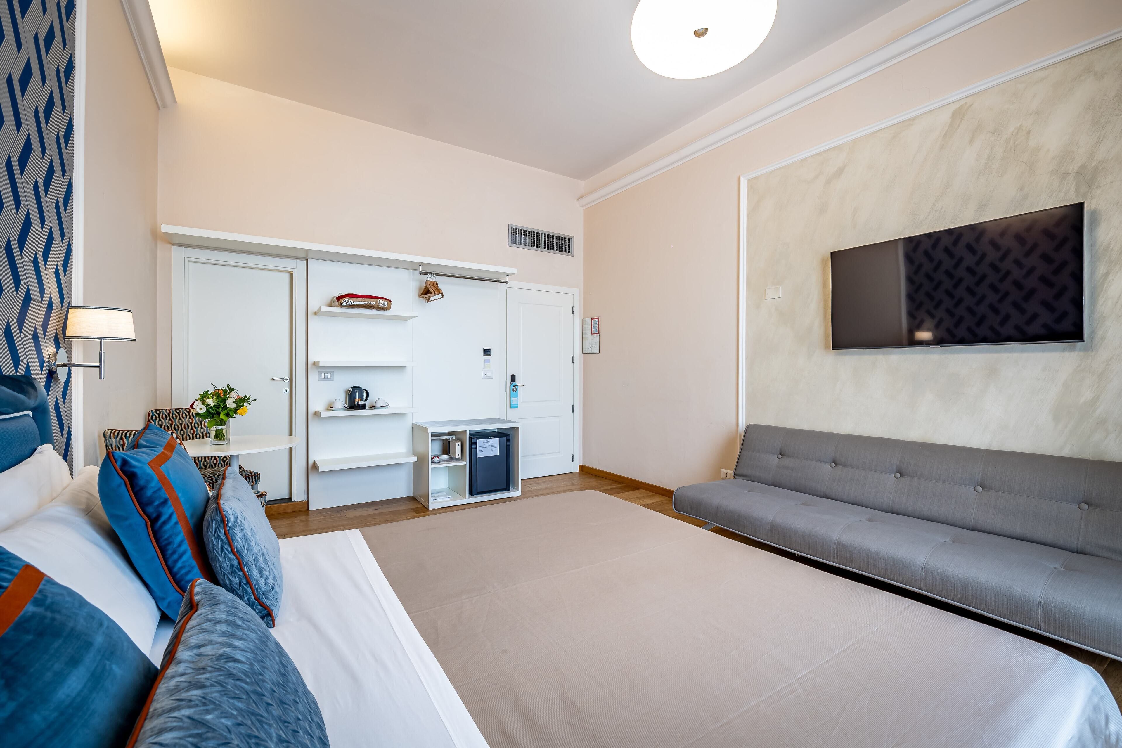 Ein modernes Hotelzimmer mit einem großen Bett, einer grauen Couch, einem flachbildfernseher an der Wand und einer kleinen Küchenzeile.