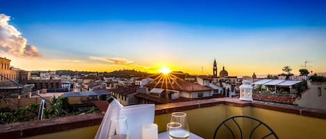Une table avec des verres à vin et une bougie, donnant sur un paysage urbain au coucher du soleil.