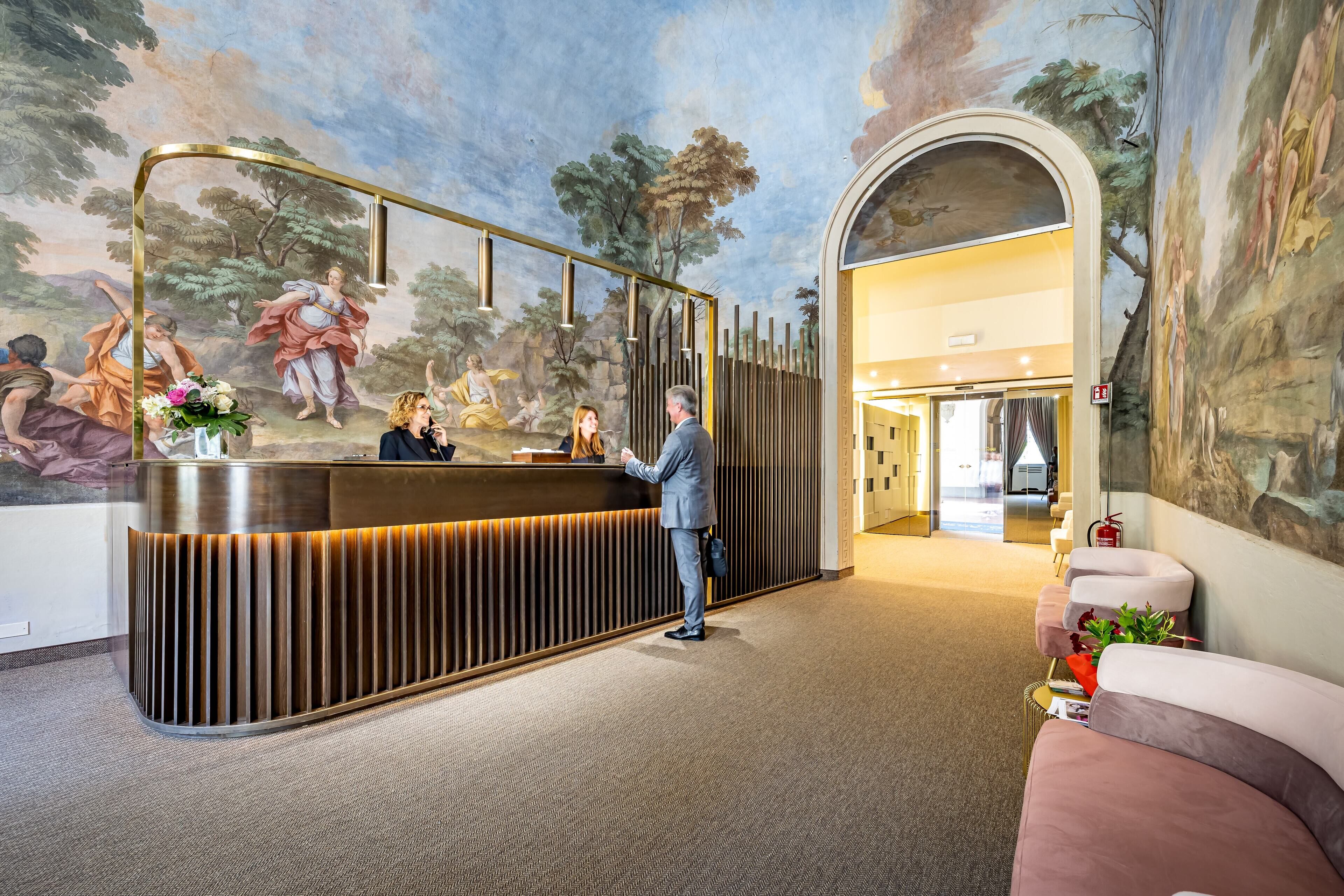 A reception desk with a modern design, a mural with classical figures, and a cozy seating area with a plant.