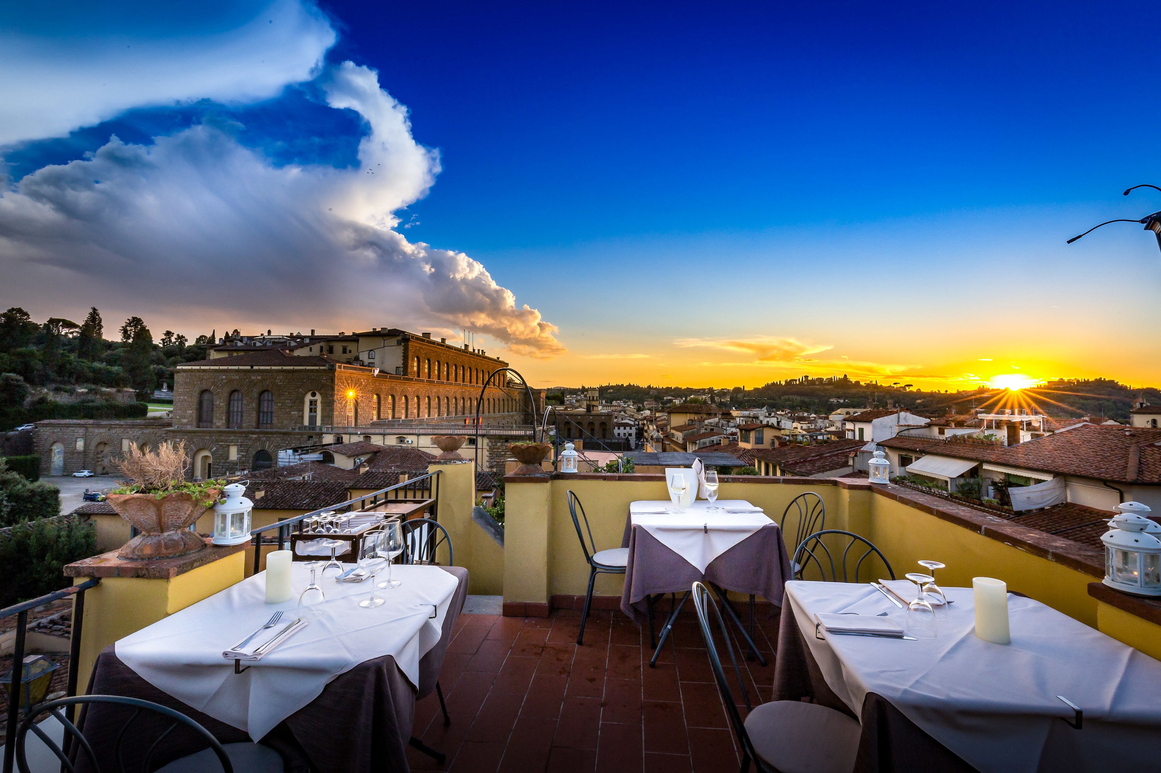 A rooftop terrace with tables set for dining, overlooking a historic town with a sunset view.