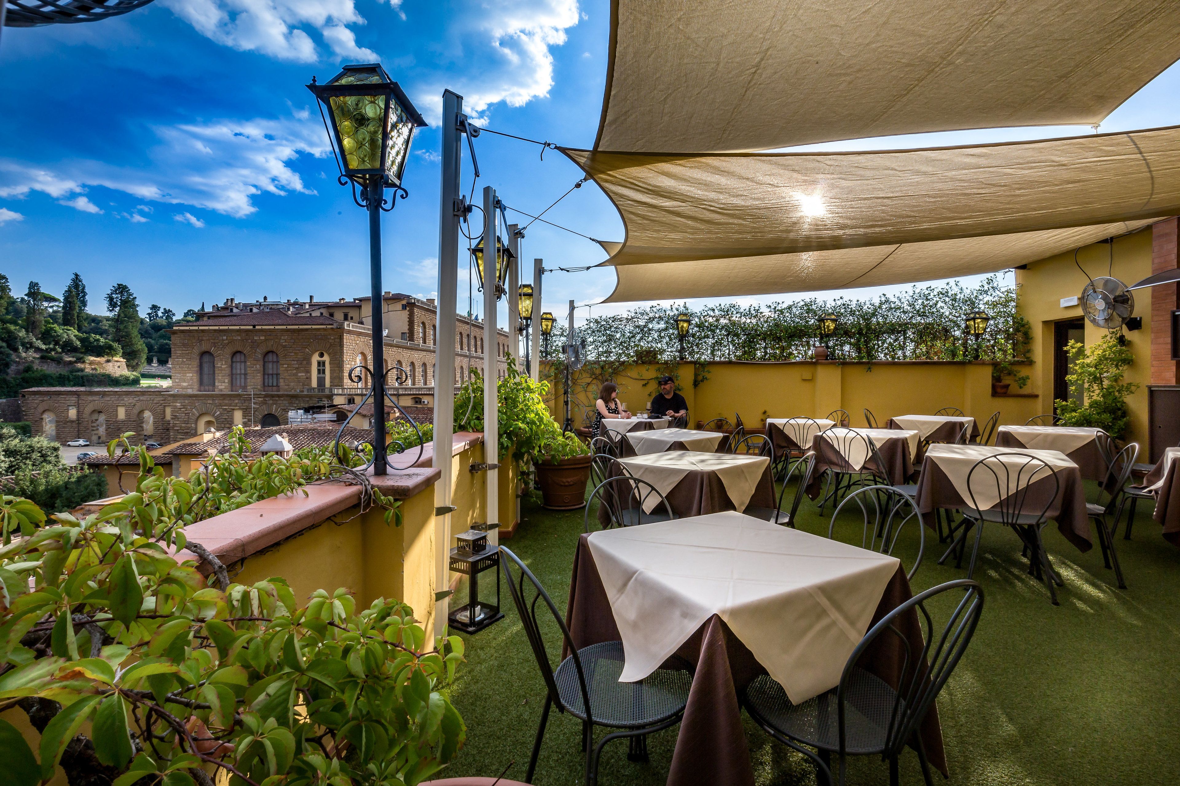 En udendørs terrasse med borde og stole, en lygtepæl og udsigt til en historisk bygning.