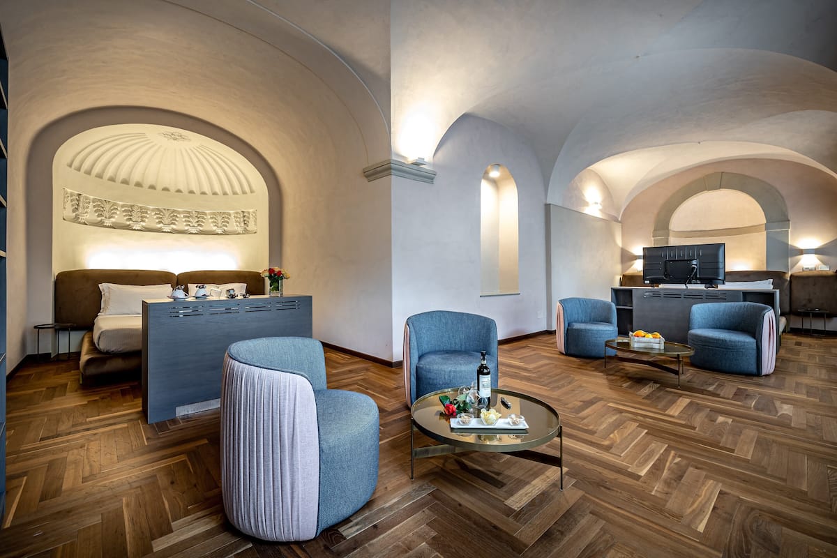 A modern hotel room with a wooden herringbone floor, a sofa, two armchairs, a small round coffee table with a bottle and flowers, and a bed with a headboard.