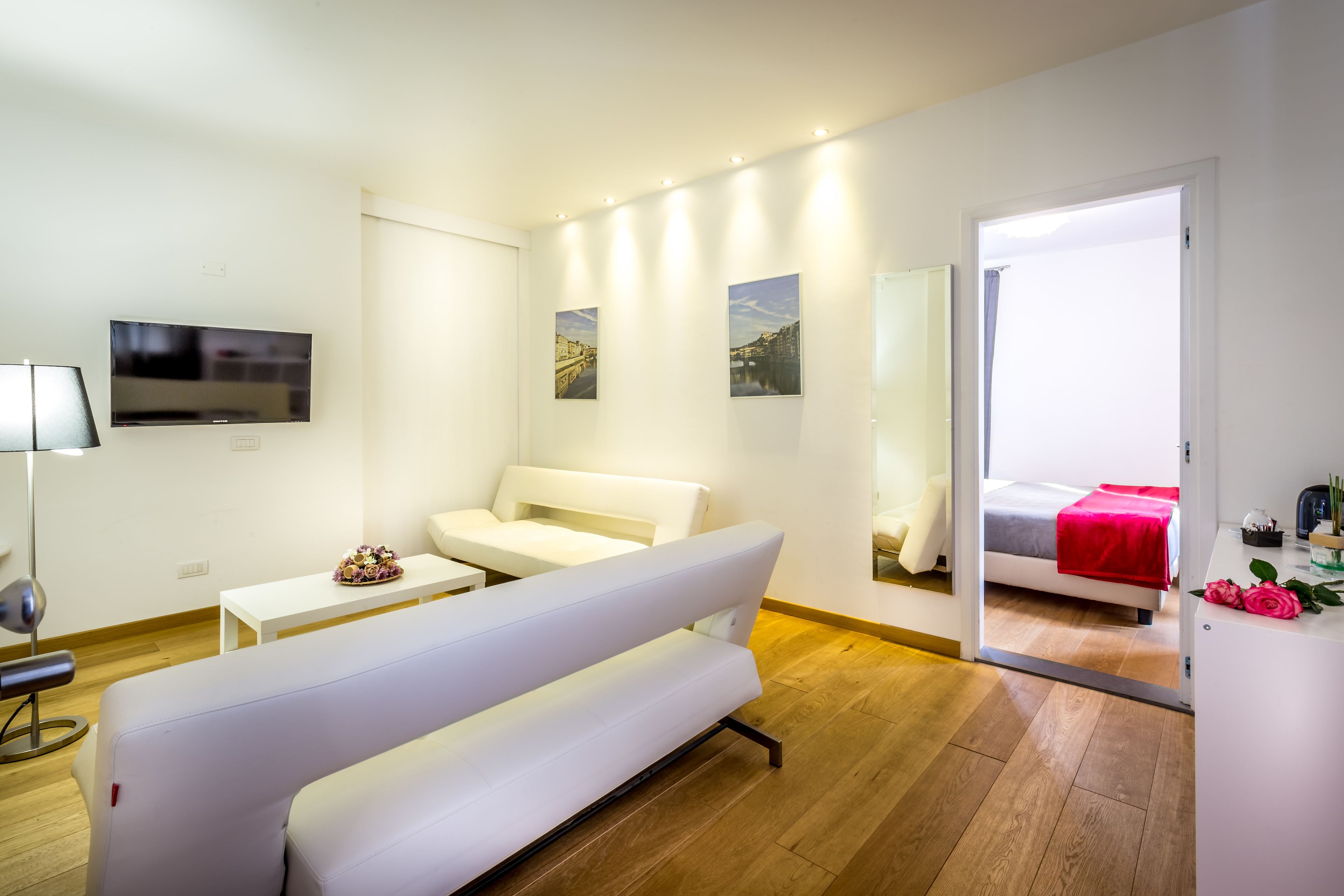 A modern hotel room with a white sofa, a flat-screen TV, a small table, and a bed with a red blanket.