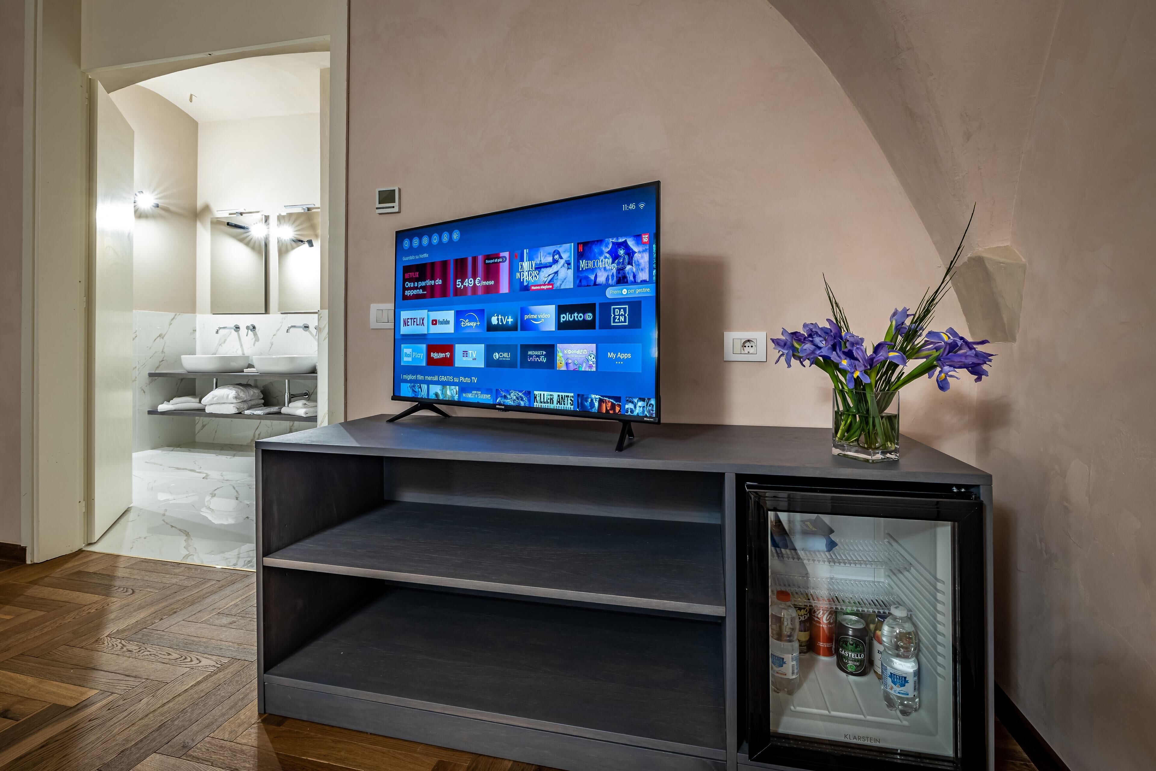 A modern living room with a flat-screen TV on a stand, a small refrigerator, and a vase with flowers.