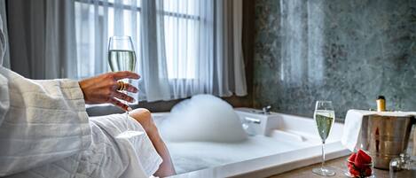 A person in a white bathrobe sitting by a bathtub with a champagne glass and a bottle of champagne.
